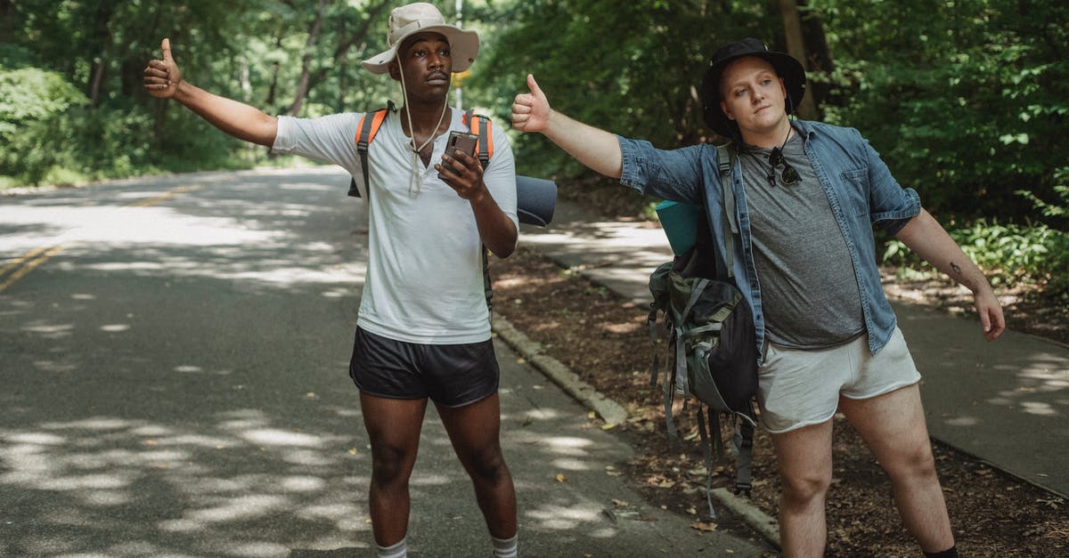 Is it possible to hitchhike to Galapagos? - Young multiethnic tourists with backpacks standing with smartphone while showing like gesture on roadway near forest and looking away