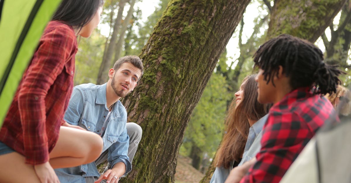 Is it safe to make groups/families sit apart from each other? - Group of young people in casual clothes sitting on plaid around near trees with moss while talking and looking at each other