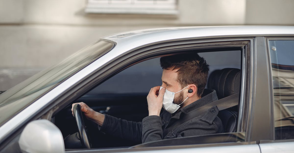 Is it safe to use earbuds in an airplane? - Serious man in disposable mask and earbuds driving car at daytime