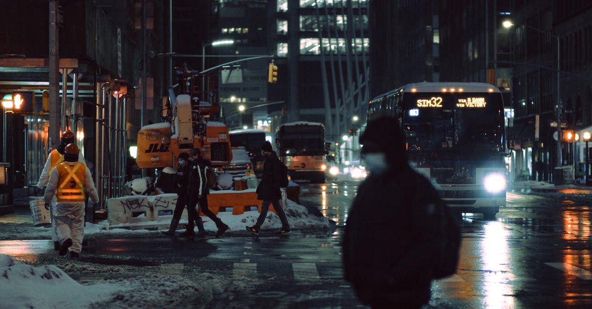 Is the late bus service from Val-d'Isère to Tignes running? - People in medical masks strolling on crosswalk near roadway with glowing buses on dark street during winter evening in city