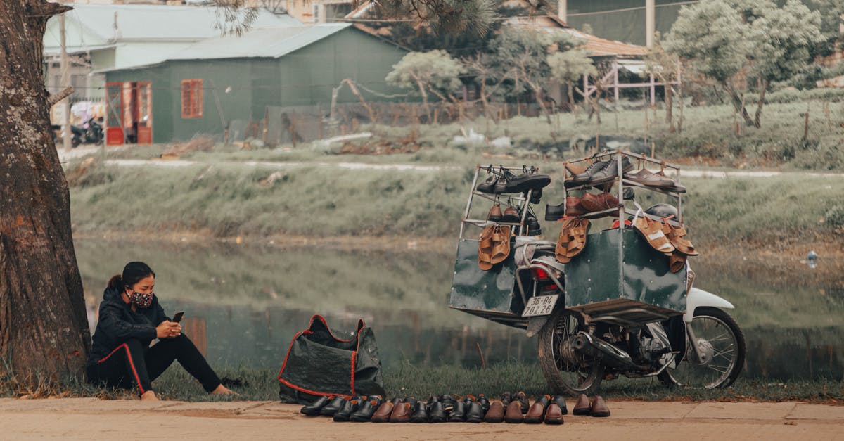 Is there a good and objective resource online about Zika virus? - Full body of focused female street seller in casual clothing and face mask surfing internet on cellphone while selling shoes by road next to pond in settlement