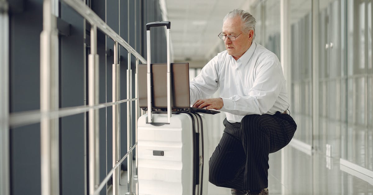 Laptop in checked baggage - Full length of focused mature male in formal wear using laptop on baggage while having semisitting position in airport corridor