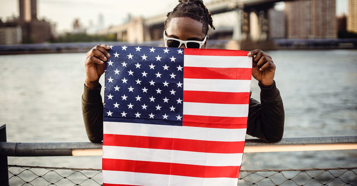 Last-minute passport for American citizens - Ethnic male in casual clothes and sunglasses standing on embankment of city river while leaning on fence showing national flag of United States of America
