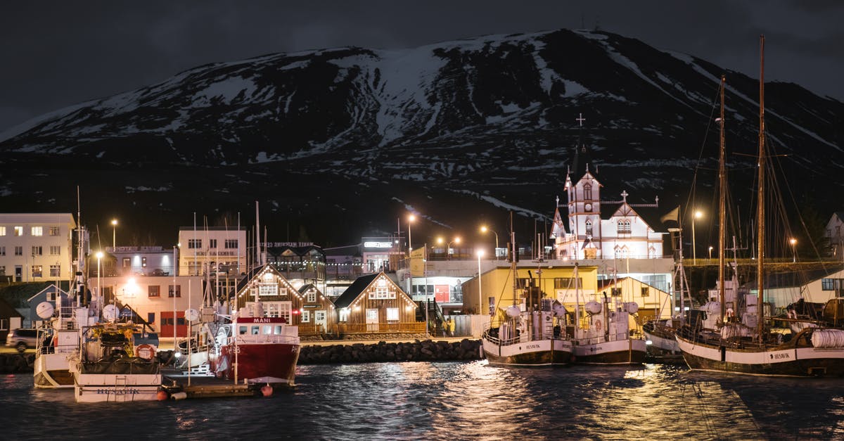 Late night travel on a Sunday in Barcelona - Seafront of brightly lit Husavik harbor late at night in Iceland with sailboats at quayside and dark snow covered mountain in background