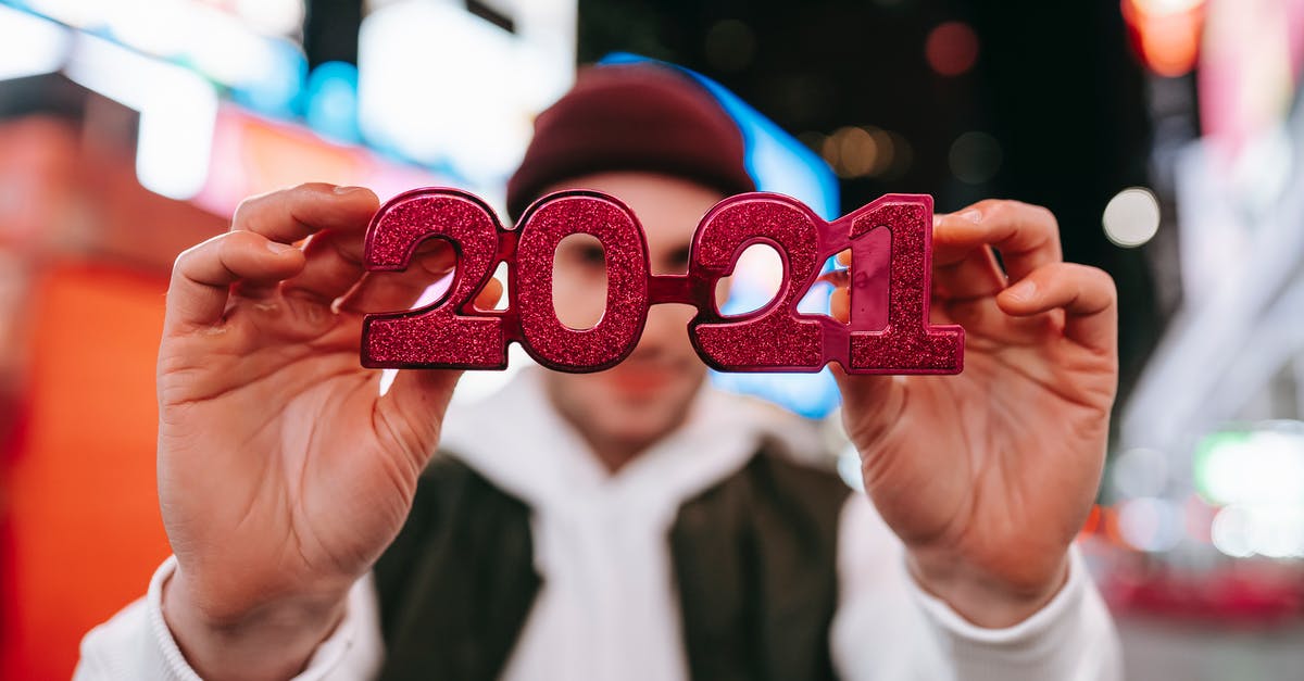 Late no-show at hotel - Anonymous male in hat demonstrating red new years 2021 glasses while standing on street with blurred signboards on modern buildings at night time