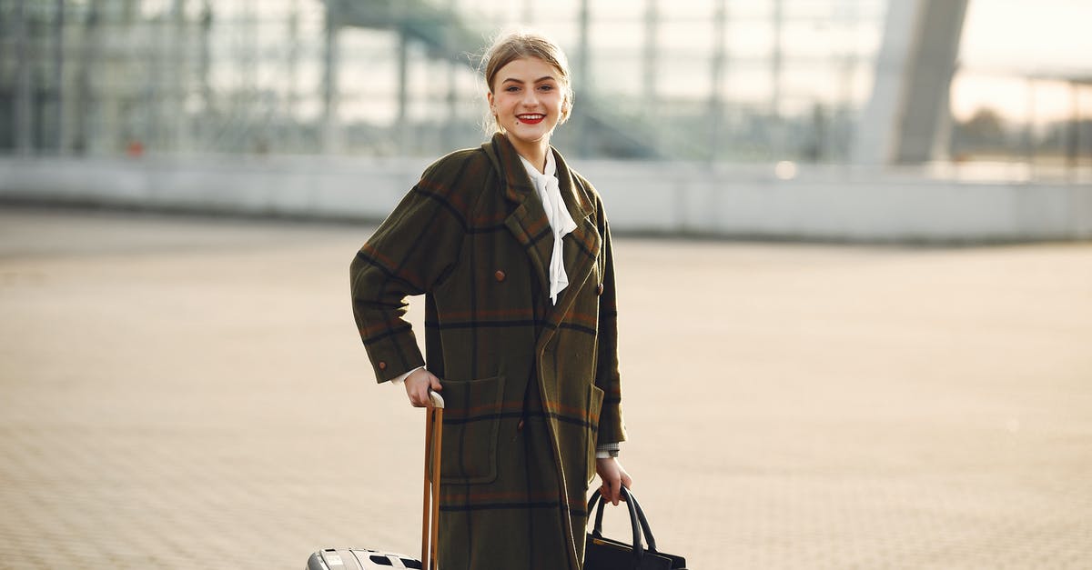 Lay over at El Prat Barcelona, luggage and city tour - Positive smiling young female traveler in casual warm coat with suitcase and ladies bag standing near airport terminal and looking at camera