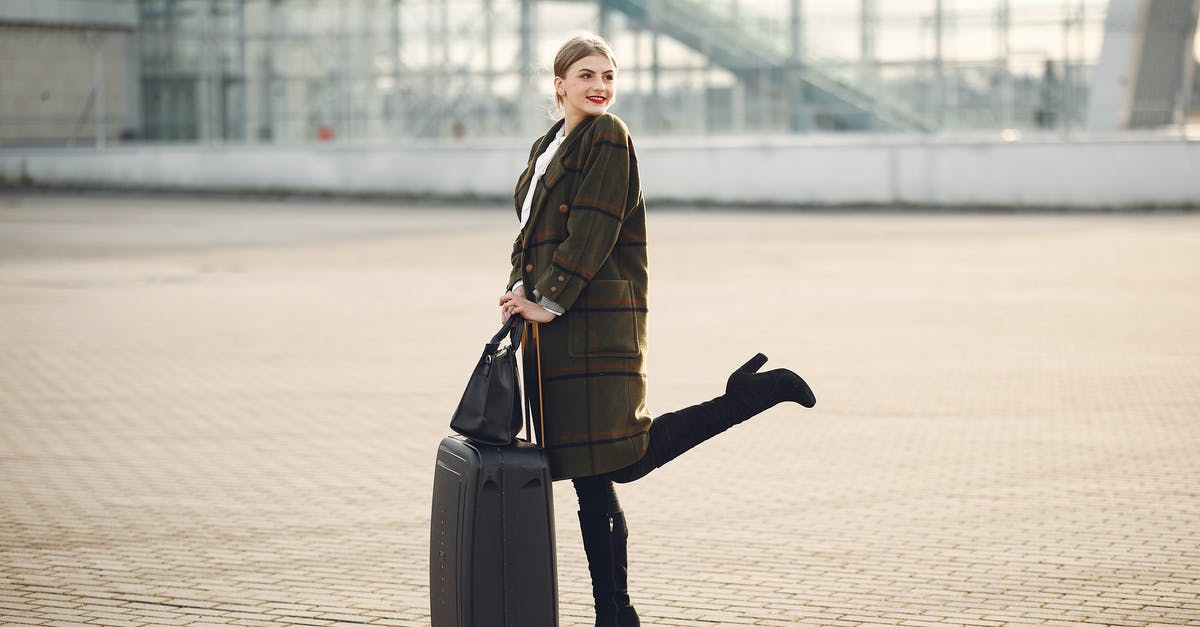 Lay over at El Prat Barcelona, luggage and city tour - Full body of stylish female tourist in warm clothes and high heeled boots standing on background of blurred airport terminal with luggage and ladies bag