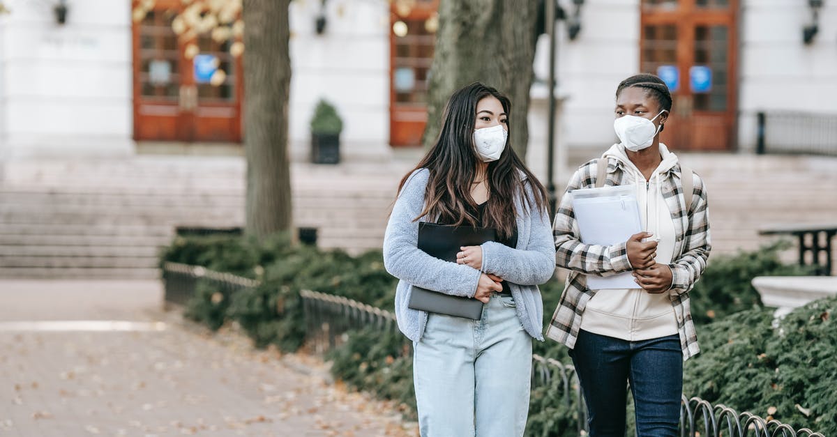 Leaving major cities during COVID-19 pandemic - Calm young multiracial female students with folders wearing casual clothes and face masks strolling together on campus sidewalk