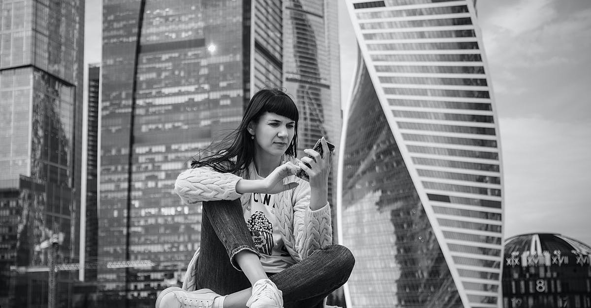 Leaving Russia to Schengen - which Ukrainian passport to use? - Black and white of young serious lady wearing casual outfit browsing smartphone while sitting on concrete fence against modern skyscrapers in Moscow Russia