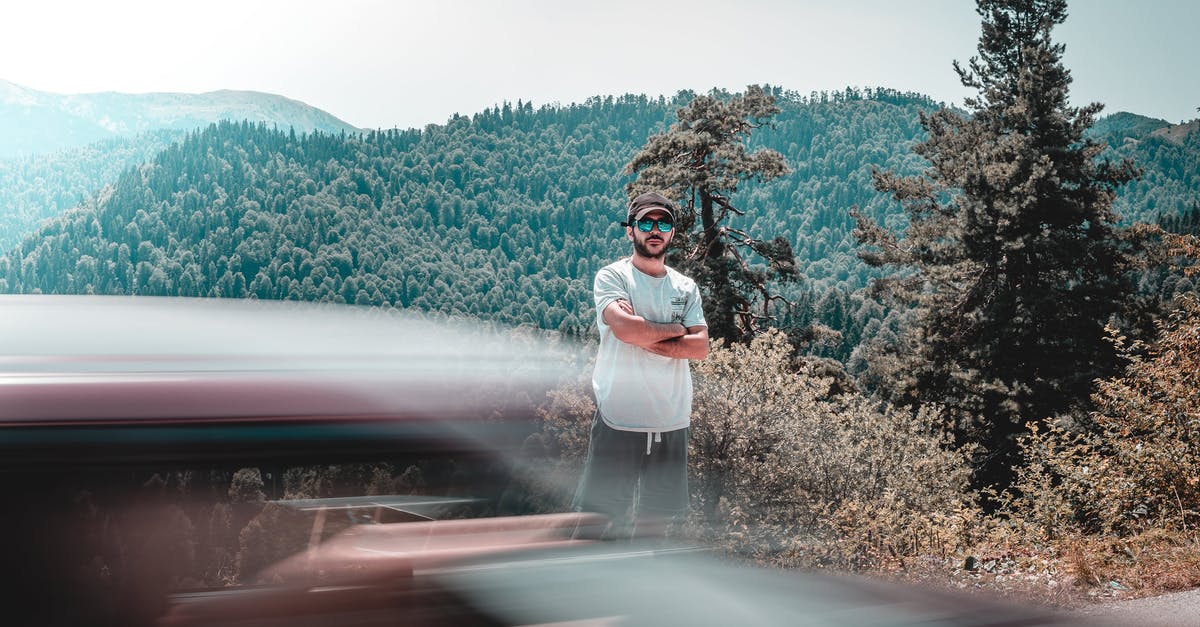 Long distance (16 hours) car trip with rental car and driver - Long exposure of male in casual clothing standing between mountains covered with woodland and car riding across road