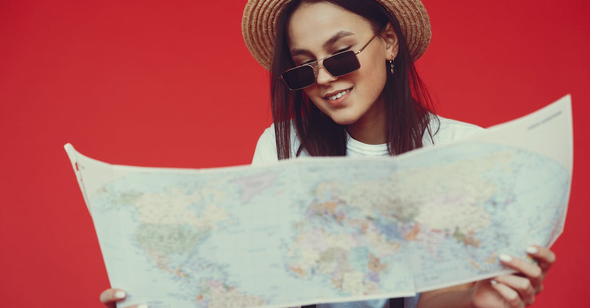 Long layover in Heathrow - with checked baggage - Smiling young woman in stylish hat and sunglasses choosing destination on paper map while standing with luggage on red background