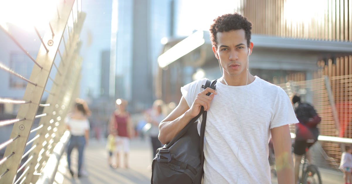 Luggage in multi-connection intercontinental air travel - Cool confident African American male in earbuds and casual clothes looking at camera with interest while standing on street with large black bag on shoulder and listening to music against blurred people and urban environment in city