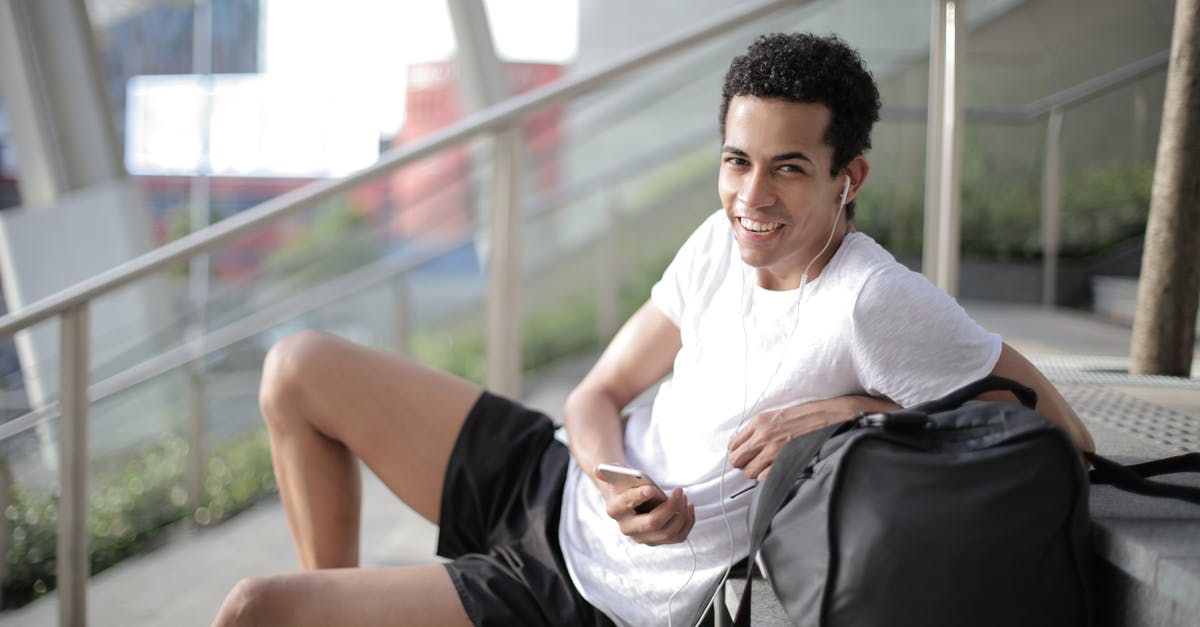 Luggage use of someone absent - Delighted African American man in earphones and sportswear smiling at camera while sitting with big black bag on stairs and listening to music against blurred exterior of contemporary building in city