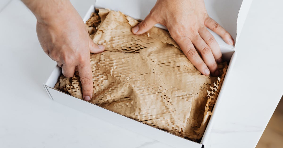 Mailing package at Heathrow during layover - From above crop adult male packing ordered product or gift in white cardboard box with craft paper for safety postal delivery at white marble table