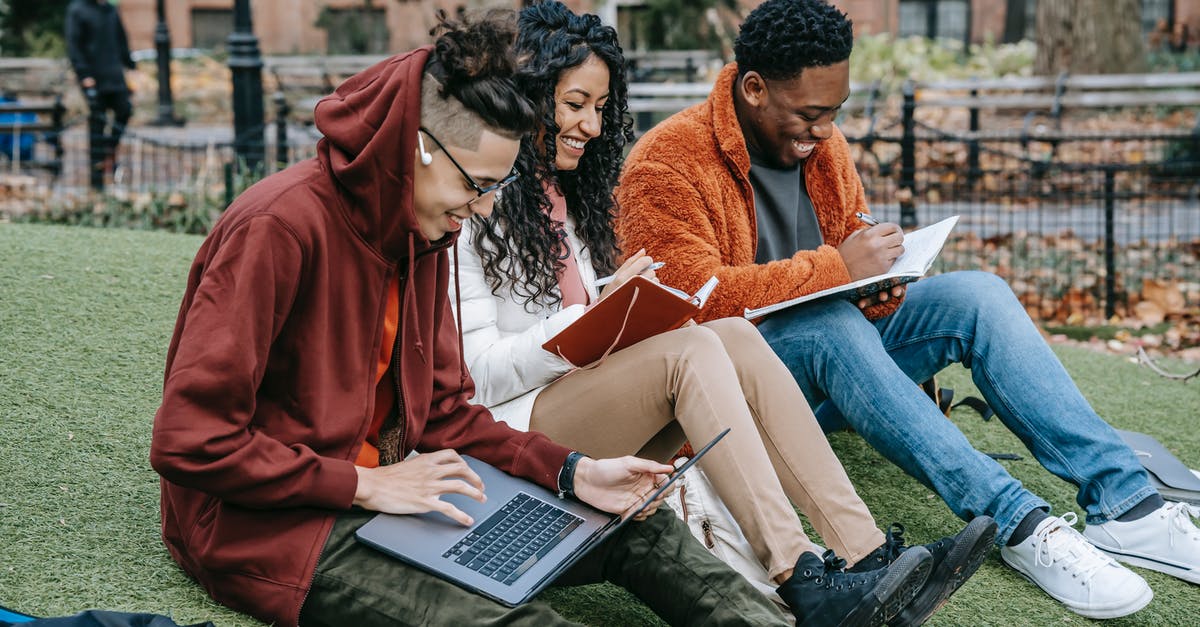 Meeting a friend at Heathrow - Happy multiracial group of students in casual clothes sitting together on grass and studying with laptop and books