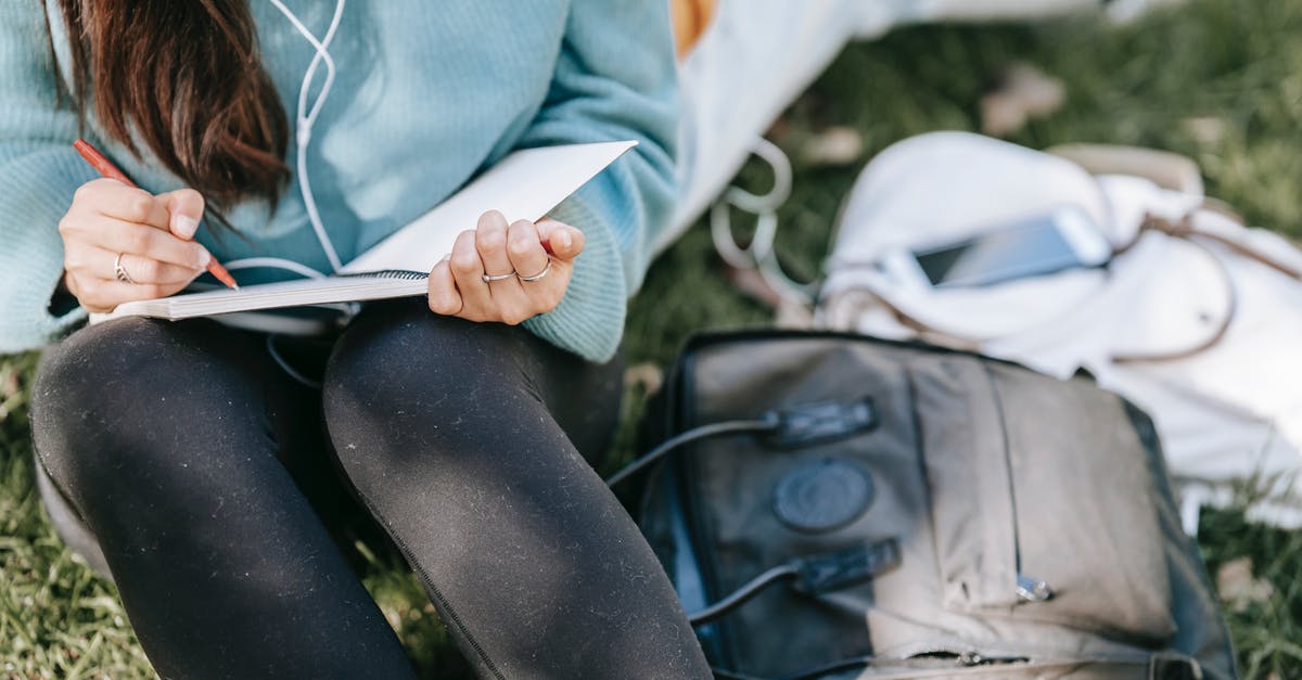 Missing bag report swindle - Crop anonymous female listening to music in earphones while writing in notebook near bags on grass