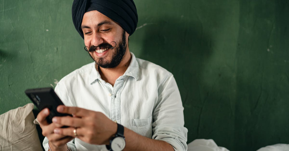 Mobile Internet Carriers in Eastern Europe [closed] - Young Indian male with black beard and mustache in casual shirt and turban messaging on smartphone and smiling happily while sitting on bed near green wall at home