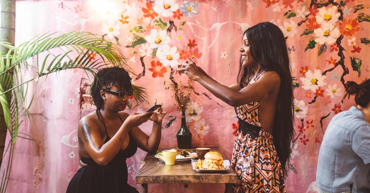 Mobile networks in Poland - Side view happy African American females in stylish dresses taking photos on smartphones of freshly cooked food while having lunch in cozy cafe against painted wall