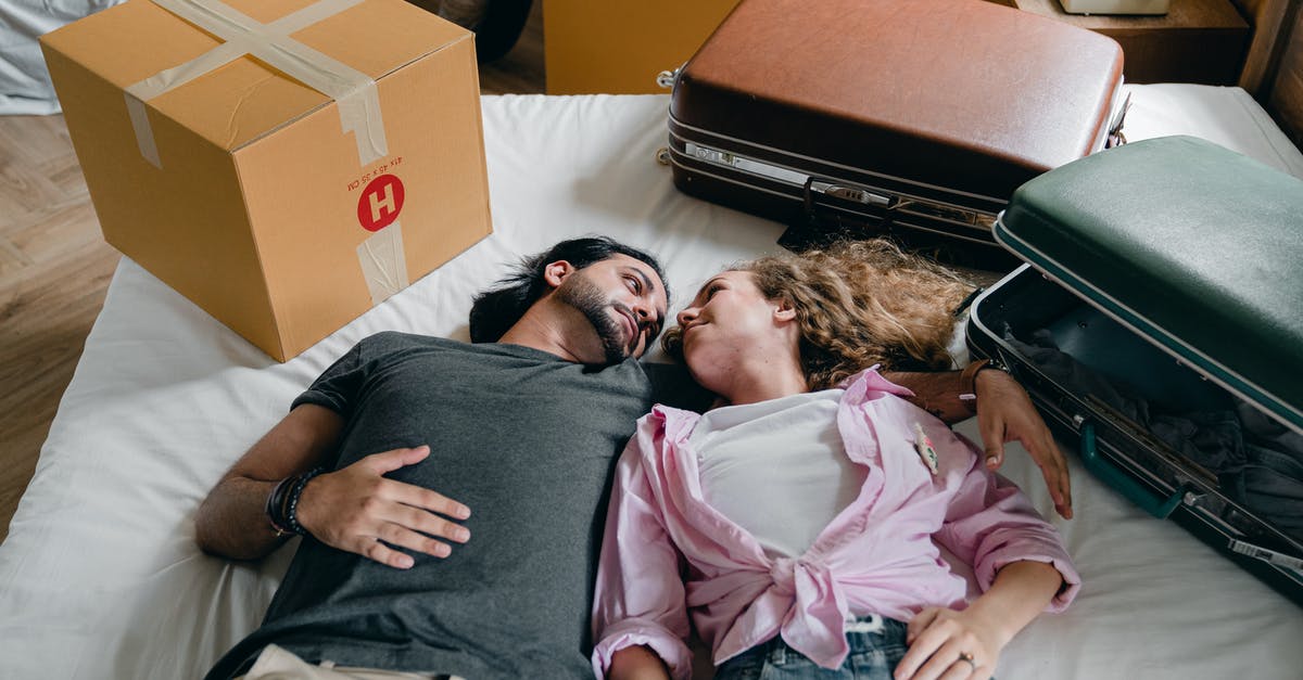 Mobile phones in luggage at Heathrow [closed] - From above of young ethnic bearded man and woman with curly hair looking at each other and lying on bed among suitcases and cardboard boxes with stuff while moving in new apartment together