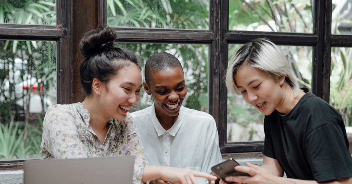 Mobile Roaming Charges in Europe - have they been abolished yet? - Cheerful young diverse female friends laughing while watching funny video on smartphone during coffee break in cozy cafe