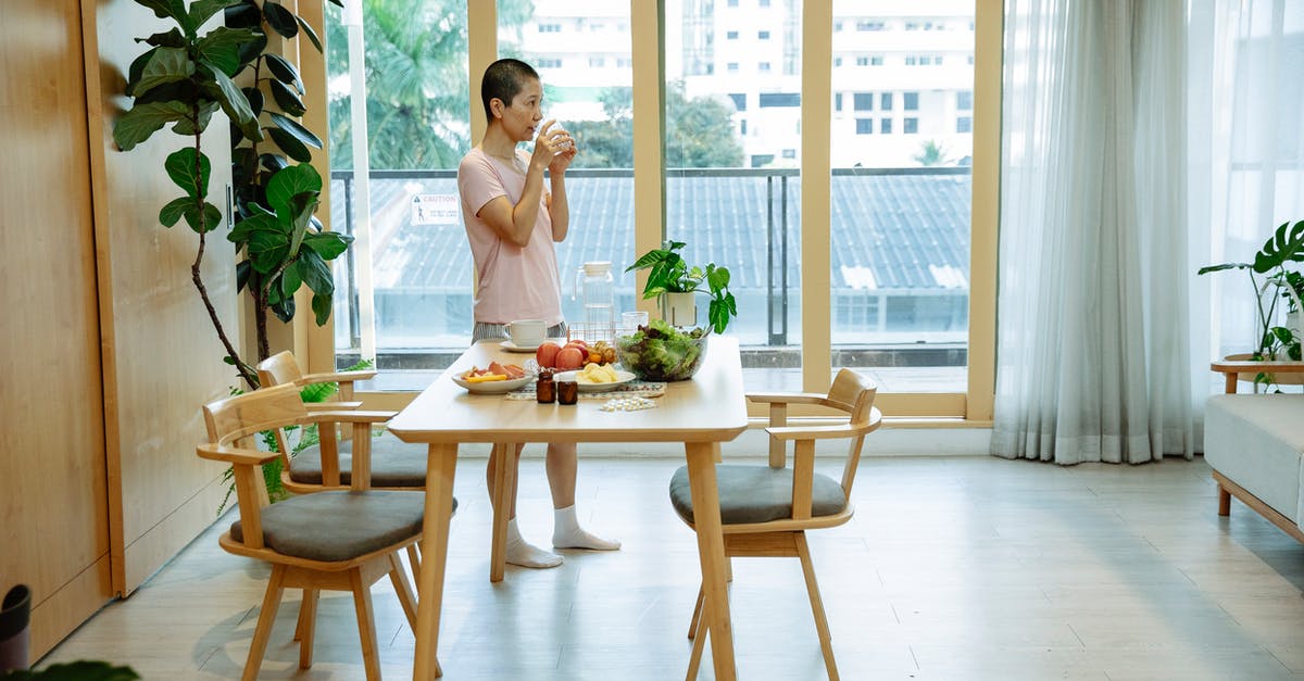 Morning-after pill availability in Poland - Side view of Asian woman with dark short hair in casual clothes drinking glass of water while standing near table with various fruits and medicines during recovery at home