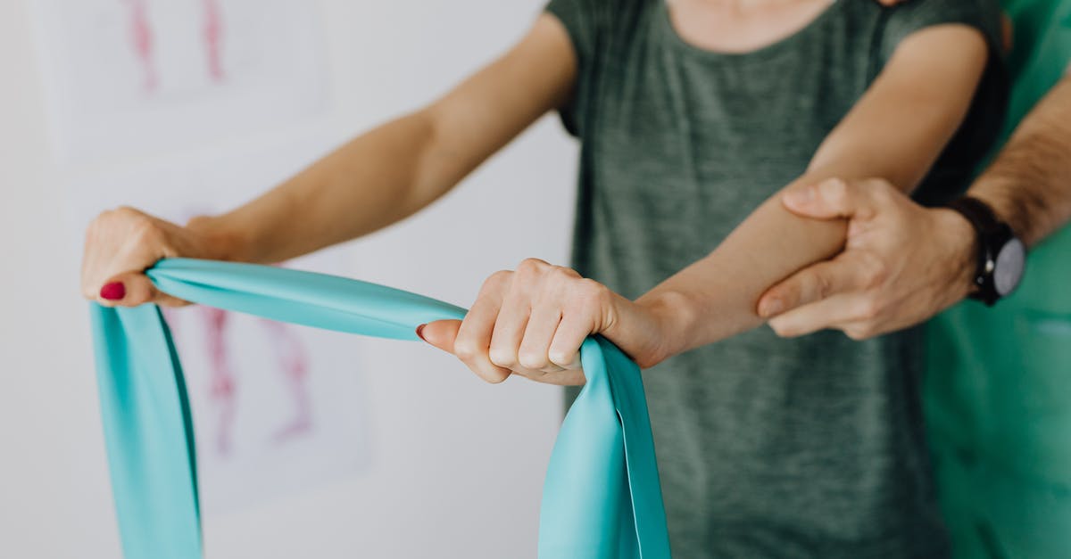 Notify border control on additional visits - Crop unrecognizable orthopedist in uniform and wristwatch supporting shoulder and reached arm of faceless female patient with resistance band in doctor office