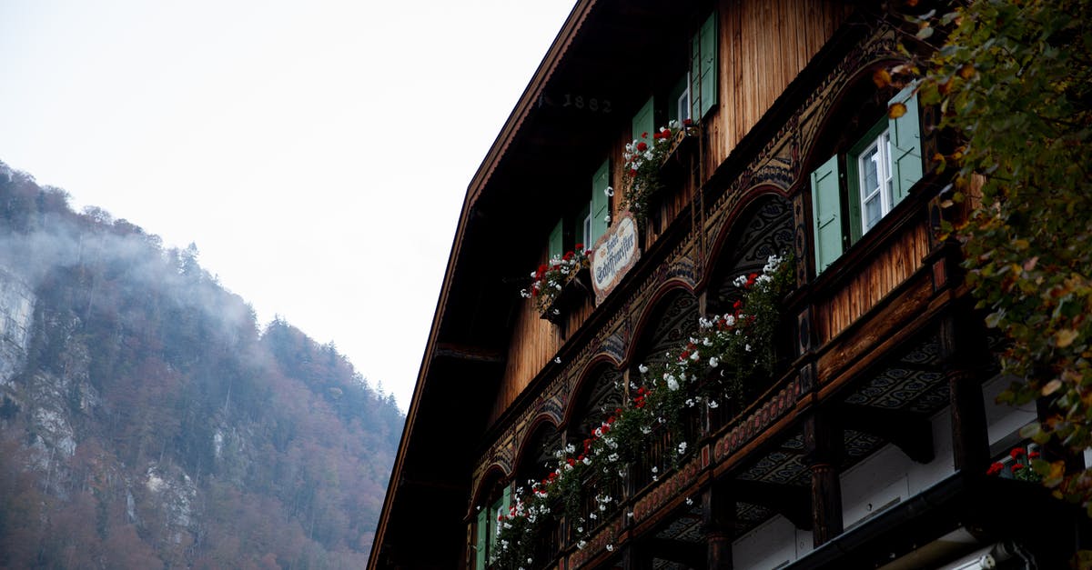 NRI Travelling to Europe from Home Country [closed] - Facade of traditional wooden house decorated with colorful potted flowers located in quiet valley with forested mountains in cloudy weather