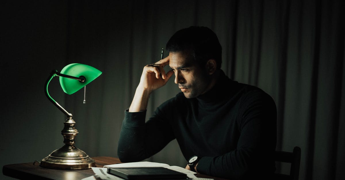 Occupancy rate and late reservations - Side view of pensive male with pen doing paperwork while sitting at wooden desktop with vintage table lamp during work in obscure room