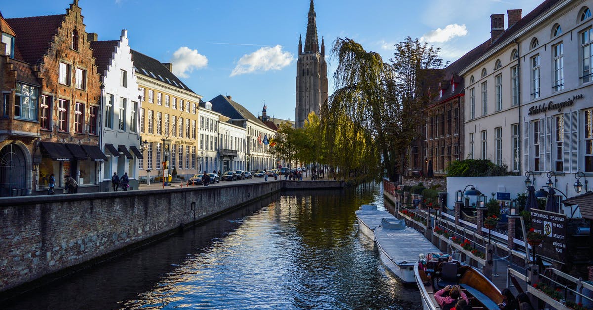 On a trip to Belgium, should I stay in Bruges or Ghent? [closed] - Picturesque scenery of rippling canal flowing near aged typical houses and historical Church of Our Lady against blue sky in Bruges