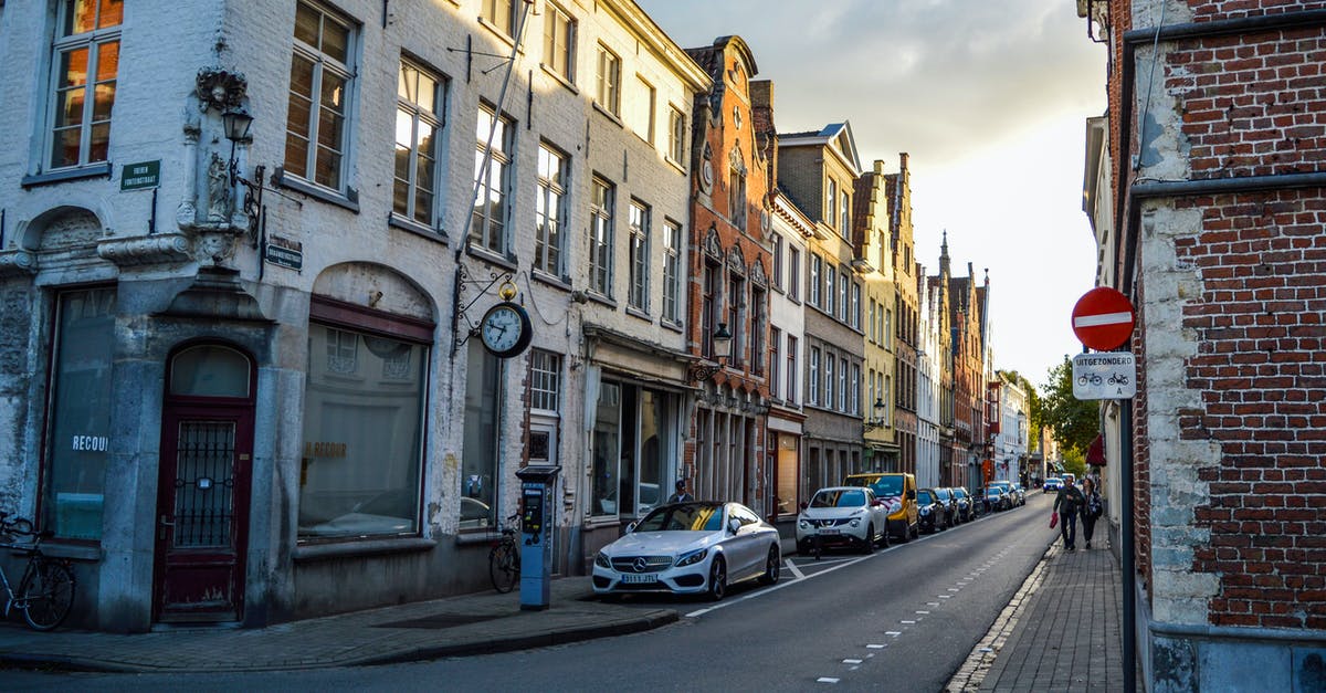 On a trip to Belgium, should I stay in Bruges or Ghent? [closed] - Low angle of asphalt road with parked vehicles near aged residential buildings in historical district of Bruges in sunny morning