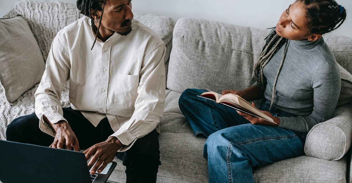 Online flight tickets booking for others - charge additional booking fee - From above of serious young African American husband and wife in casual outfits sitting on sofa with book and laptop and discussing relationships in cozy living room