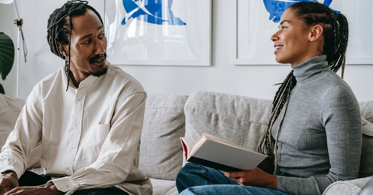 Online flight tickets booking for others - charge additional booking fee - Positive young black lady with Afro braids sitting on sofa with interesting book in hands and speaking with boyfriend working remotely on laptop