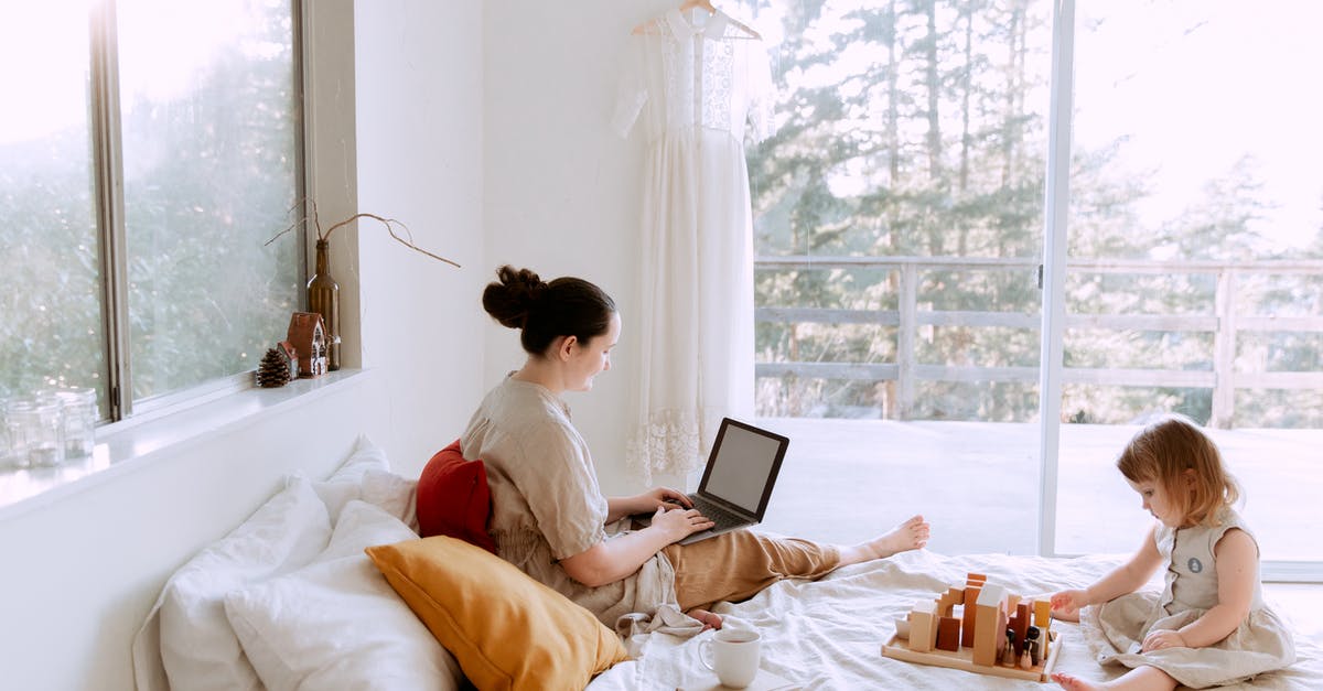 Online resource for duty-free - Side view of cute toddler girl sitting on bed barefoot and playing with colorful wooden blocks while mother using laptop in bed enjoying morning coffee