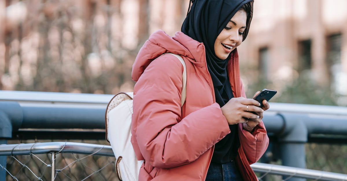 Online ticketing of National Express buses in UK - Muslim ethnic female in hijab texting message on mobile phone near iron construction on blurred background of building