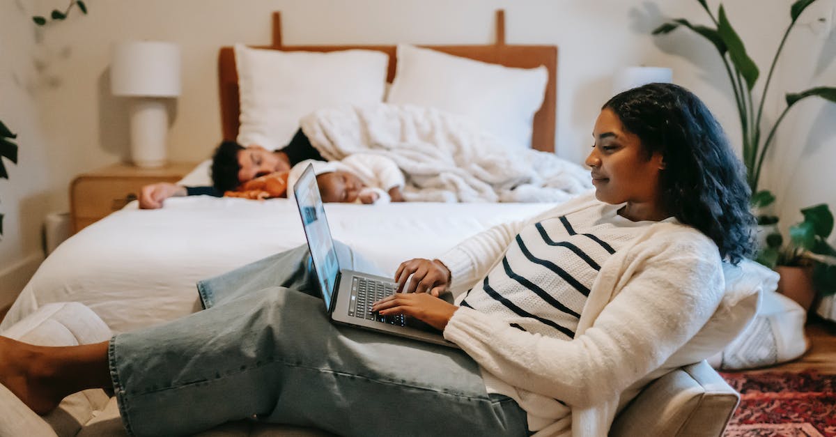 Overnight connection in Guangzhou - where do I sleep? - Side view of pensive ethnic woman freelancer sitting on couch and browsing netbook while man and little baby sleeping on bed in cozy bedroom