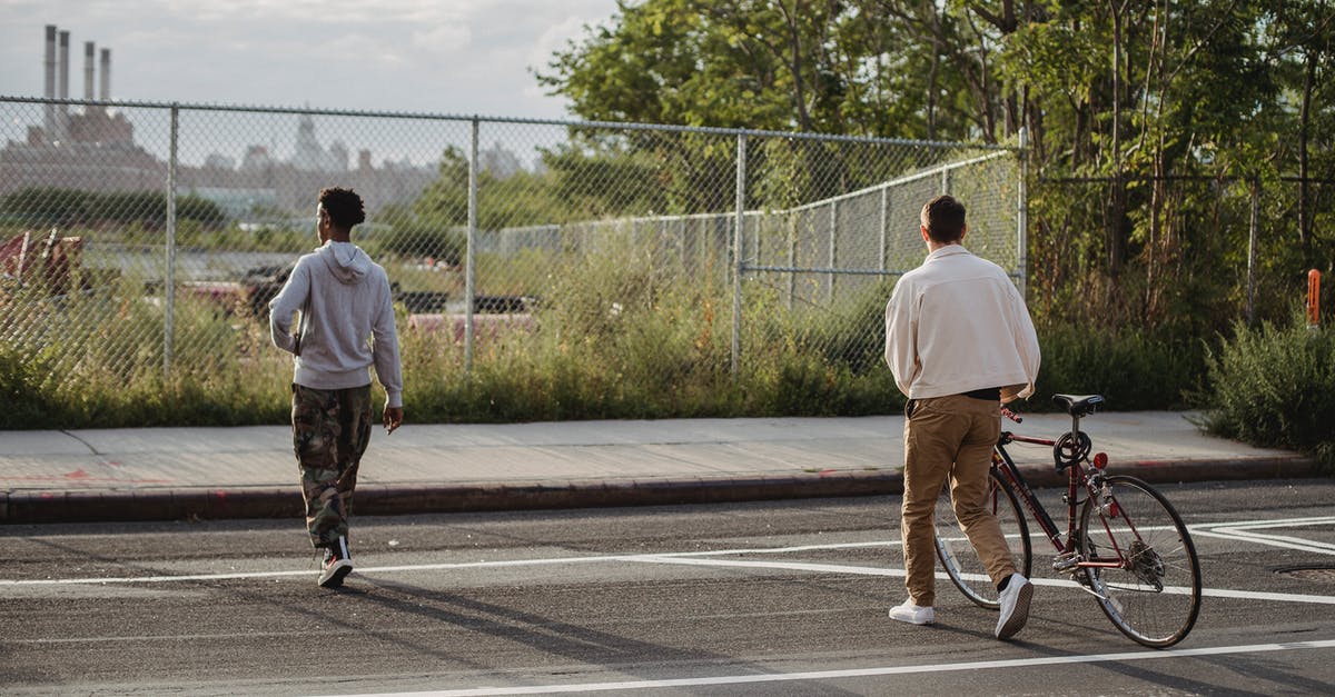 Park and Ride in Copenhagen - Back view of anonymous young male millennial in trendy outfit rolling bicycle while crossing asphalt road with African American friends in city outskirts