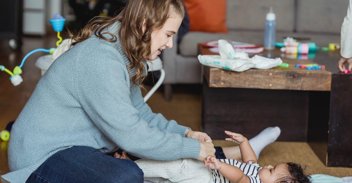 Passport queue length in UK in relation to arrival method - Full body cheerful young mother playing with and putting pants on cute ethnic baby lying on floor at home