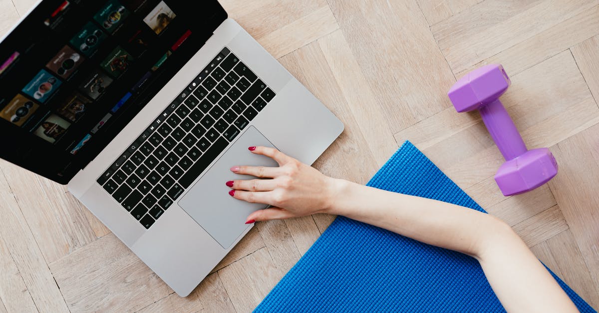 Photographable location from "Gangnam Style" music video - Top view of crop anonymous female looking for video workout courses on laptop while sitting on blue yoga mat with purple dumbbell beside on parquet floor