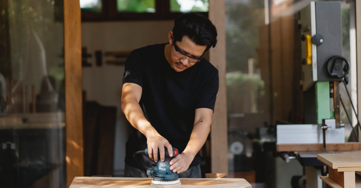 Polish Airports' fingerprint Process - Concentrated male master in protective glasses smoothing wooden plank with sander while creating wooden detail in workroom