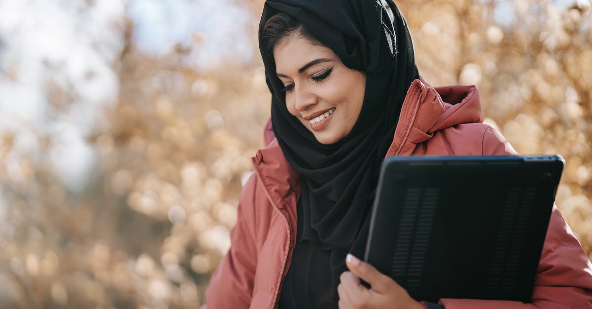 Precautions when travelling to Middle East (DXB,SHJ) w.r.t PC and Kindle contents? - Smiling Muslim woman freelancer in black headscarf standing with netbook in hand and looking down on street against tree with autumn foliage in daylight