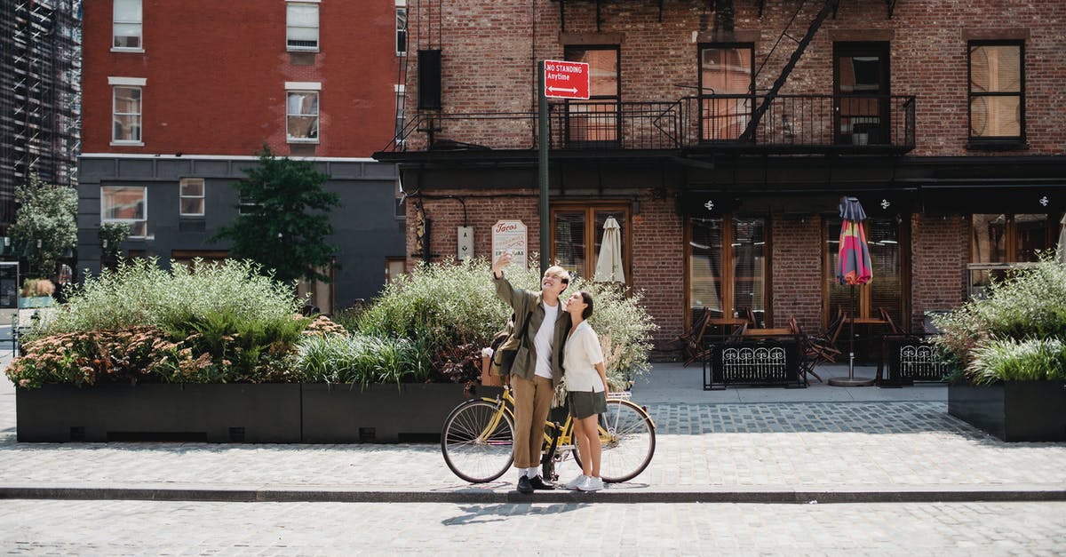 Present for bicycle traveler [closed] - Full body of anonymous joyful young tourists standing on paved sidewalk near aged buildings and parked bicycle and taking self portrait on mobile phone