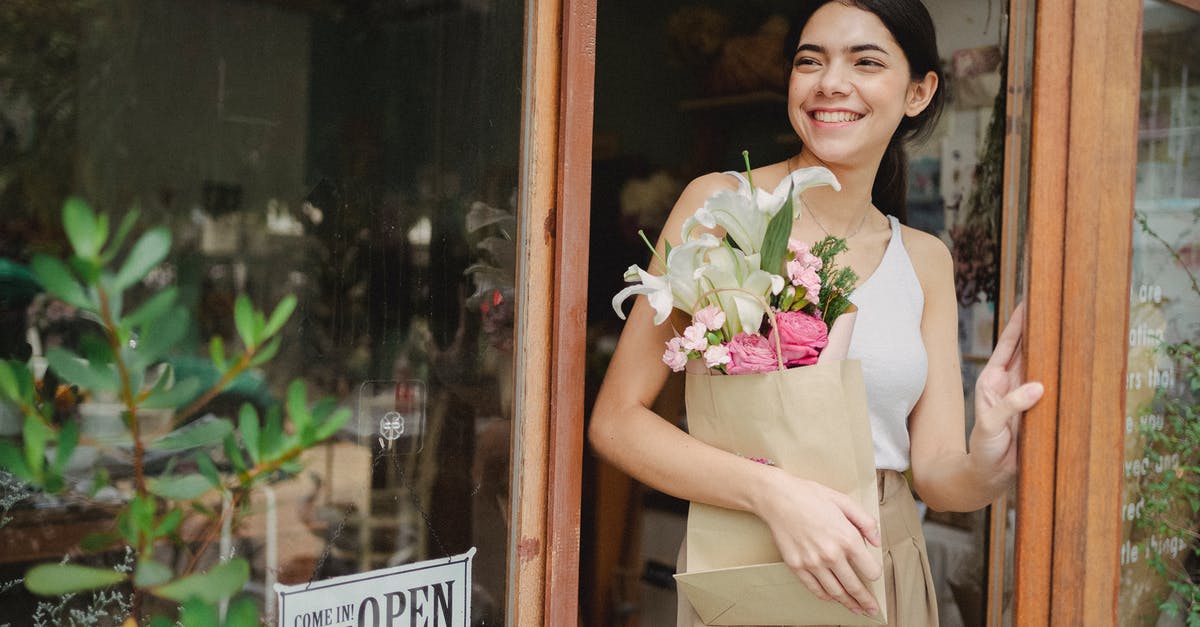 Purchase extra baggage now or wait till checkin time? - Toothy smiling female standing near flower shop and holding paper package with bouquet