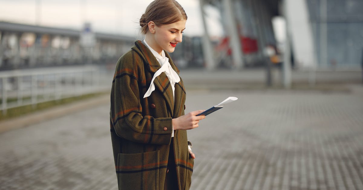 Purchasing train tickets at Helsinki airport - Smiling female passenger wearing trendy plaid coat and white blouse checking passport and ticket standing on pavement near contemporary building of airport