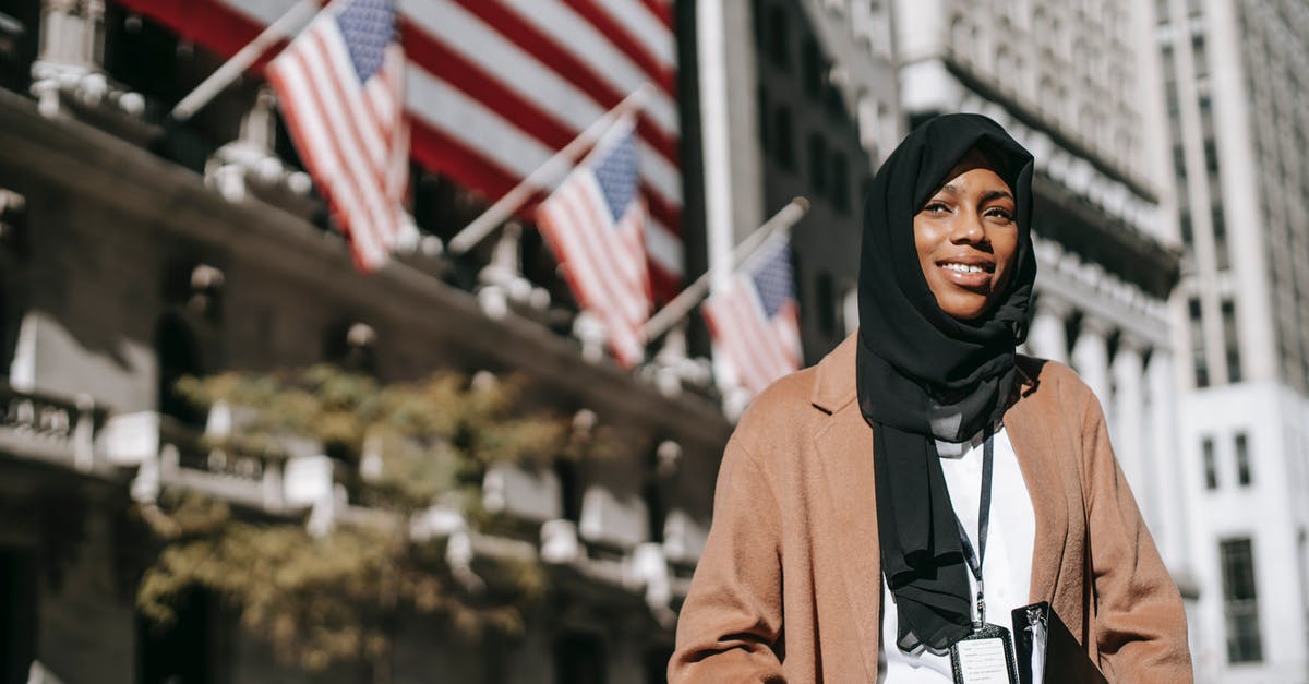 Quickly obtaining an ID to fly within the US - From below of cheerful African American female ambassador with folder wearing hijab and id card looking away while standing near building with American flags on blurred background
