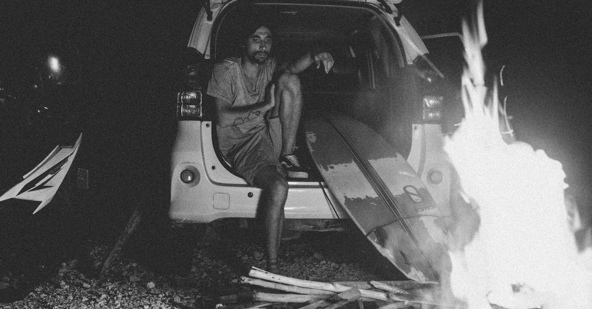 Random camping along Central and South Californian coastline - Black and white bearded young male surfer in summer clothes recreating in SUV car trunk and looking at bonfire at night