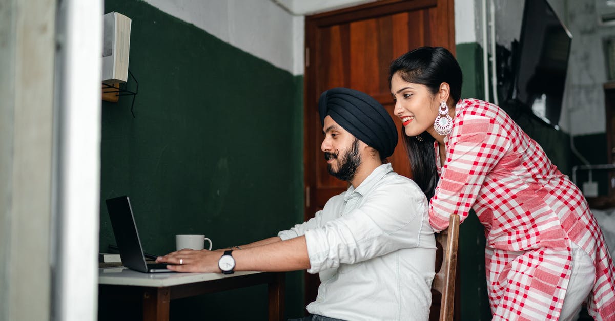 Re-entering Uk after a work visa ... please help [closed] - Side view of positive young Sikh man in shirt and turban working on laptop at home while wife leaning on chair behind