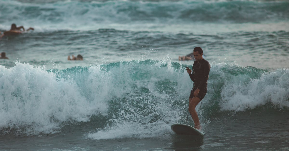 Re-entry to Germany after vacation using blue card - Young male surfer in short wetsuit riding surfboard and using mobile in magnificent restless sea with other surfers paddling behind