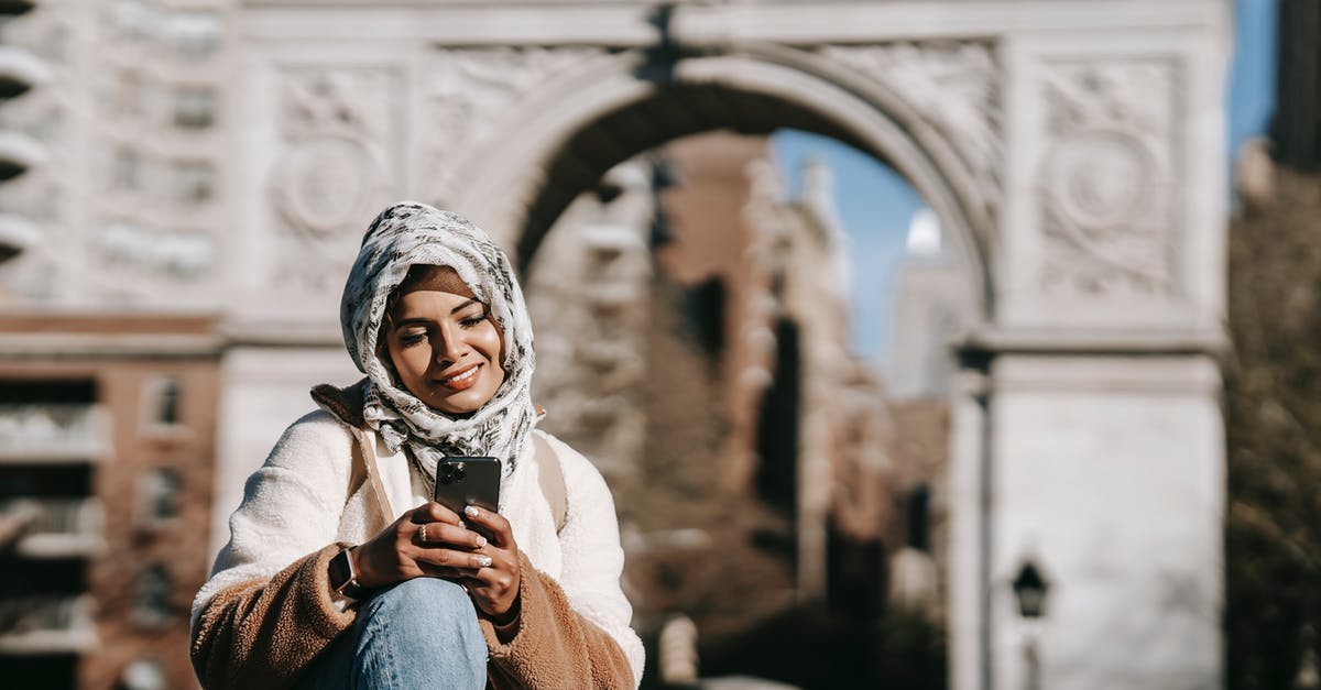 Re-entry to Germany after vacation using blue card - Self assured young ethnic female in stylish clothes and Muslim headscarf smiling while messaging on smartphone sitting in city square near aged arch