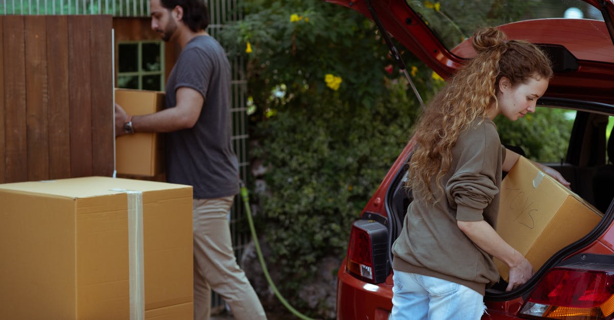 Rent car Green Card in Dubrovnik (Croatia) - Focused young woman with curly hair taking cardboard package out from red automobile trunk on street near stack of moving boxes while bearded ethnic man carrying box into new house