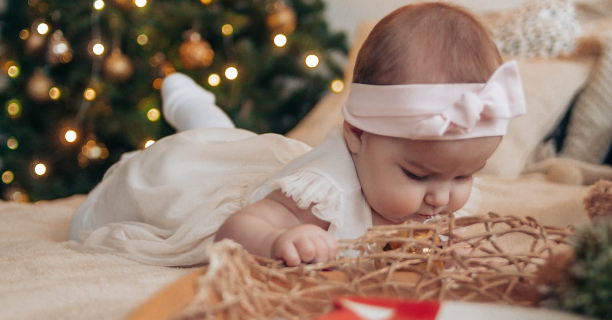 Renting a holiday house in the Riviera with a small child - Toddler girl in white dress lying on comfortable bed near pillows and decorative objects in light bedroom with Christmas tree with baubles and garland at home